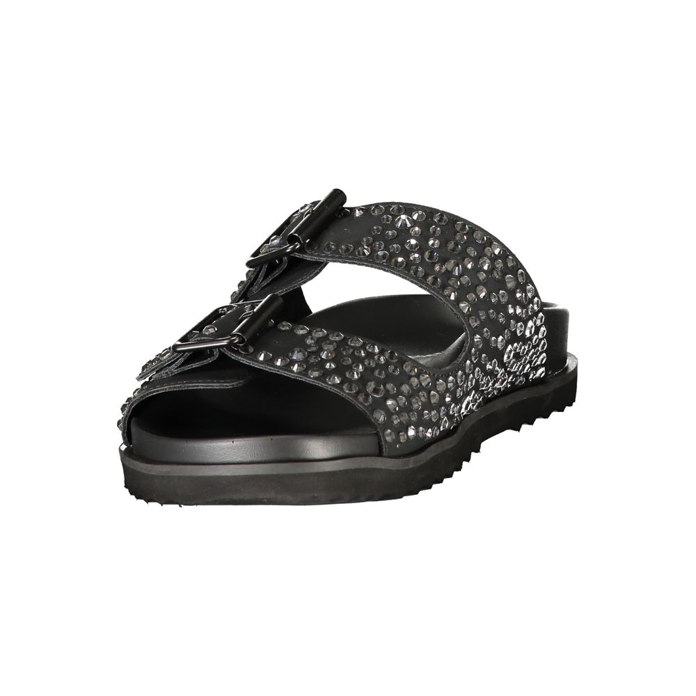 Female model wearing black double-strap slide sandal with two wide straps densely encrusted with gunmetal crystal rhinestones, dual adjustable buckle closures, and a sleek black ridged outsole, captured mid-step in a casual lifestyle pose showcasing the sandal's bold crystal-embellished glamour and effortless rock-luxe edge