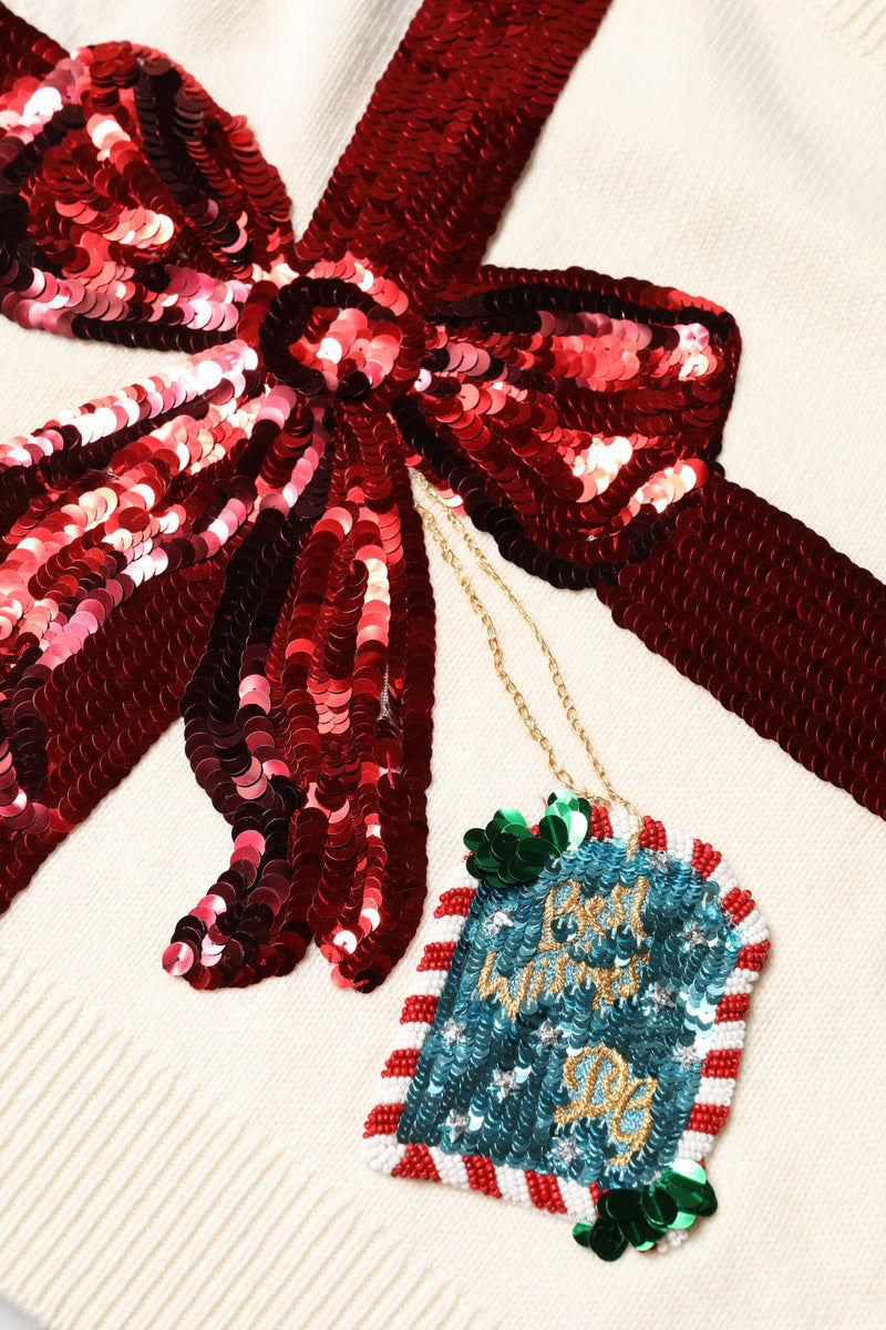 White knit crew neck sweater featuring a large red sequin bow and dangling sequin gift tag detail